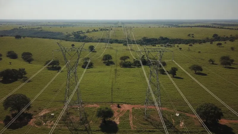 Linhas de transmissão de energia em paisagem rural do Centro-Oeste, com torres metálicas e cabos se estendendo ao horizonte sob um céu azul.